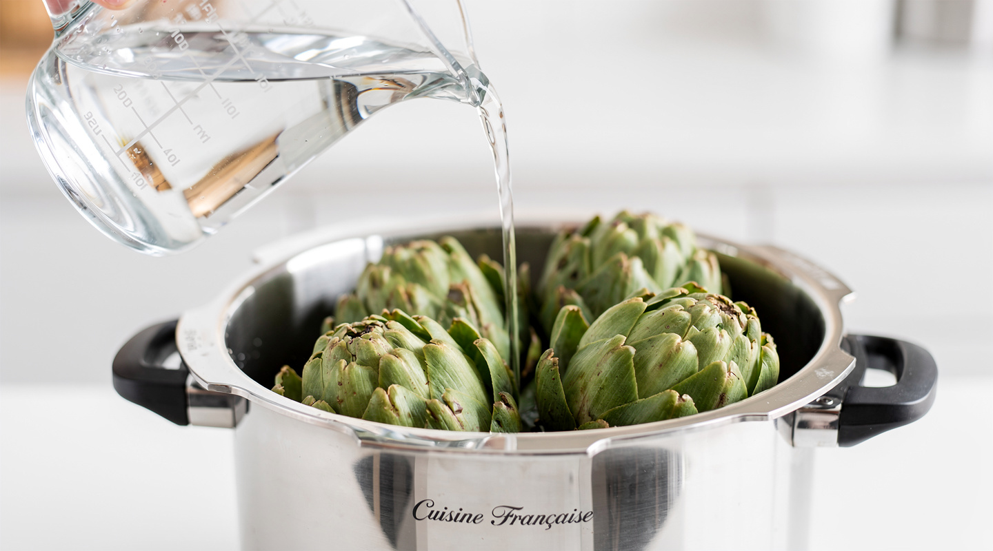 Una taza medidora vertiendo agua en una olla a presión de acero inoxidable que contiene alcachofas