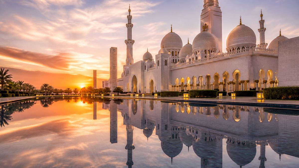 Grande mosquée blanche aux multiples dômes reflétée dans un bassin au coucher du soleil